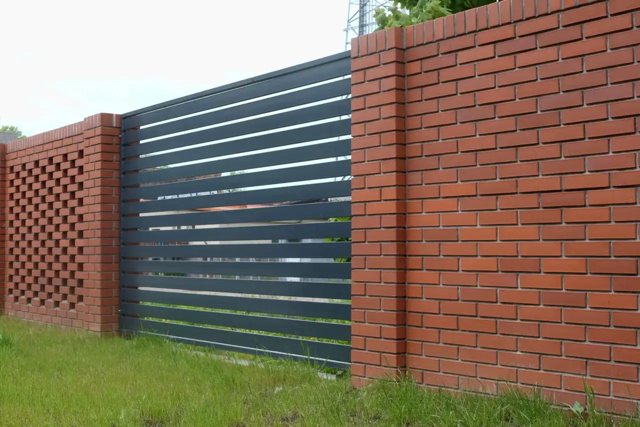 Nowoczesne ogrodzenie z cegły z ciemnoszarą, poziomą bramą. Ceglane słupki i fragment muru z ażurowym wzorem.