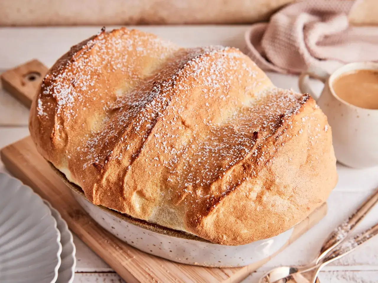Ein goldbraunes, luftiges Salzburger Nockerl, bestäubt mit Puderzucker, in einer Auflaufform. Daneben eine Tasse Kaffee und Servietten.
