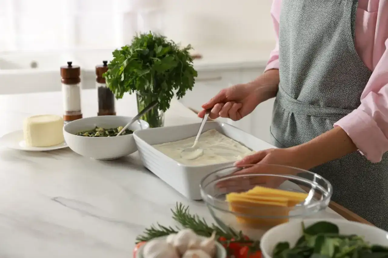 Przygotowanie sosu beszamelowego do lasagne. Na blacie widać składniki: masło, szpinak, makaron, zioła i czosnek.