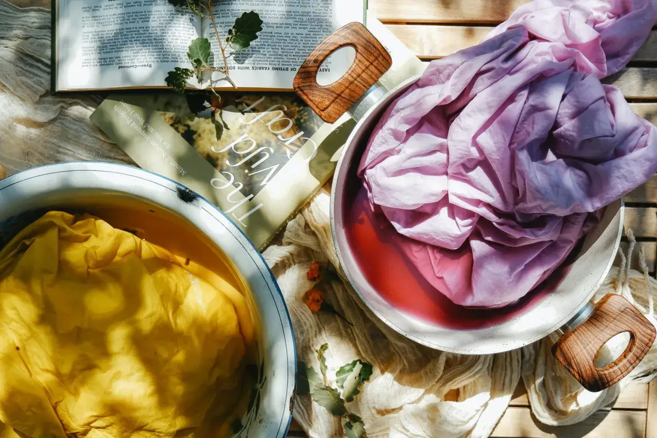 Dyeing fabrics with natural dye. Yellow fabric in a bowl, pink fabric in a pot of red liquid, and a book on natural dyeing.