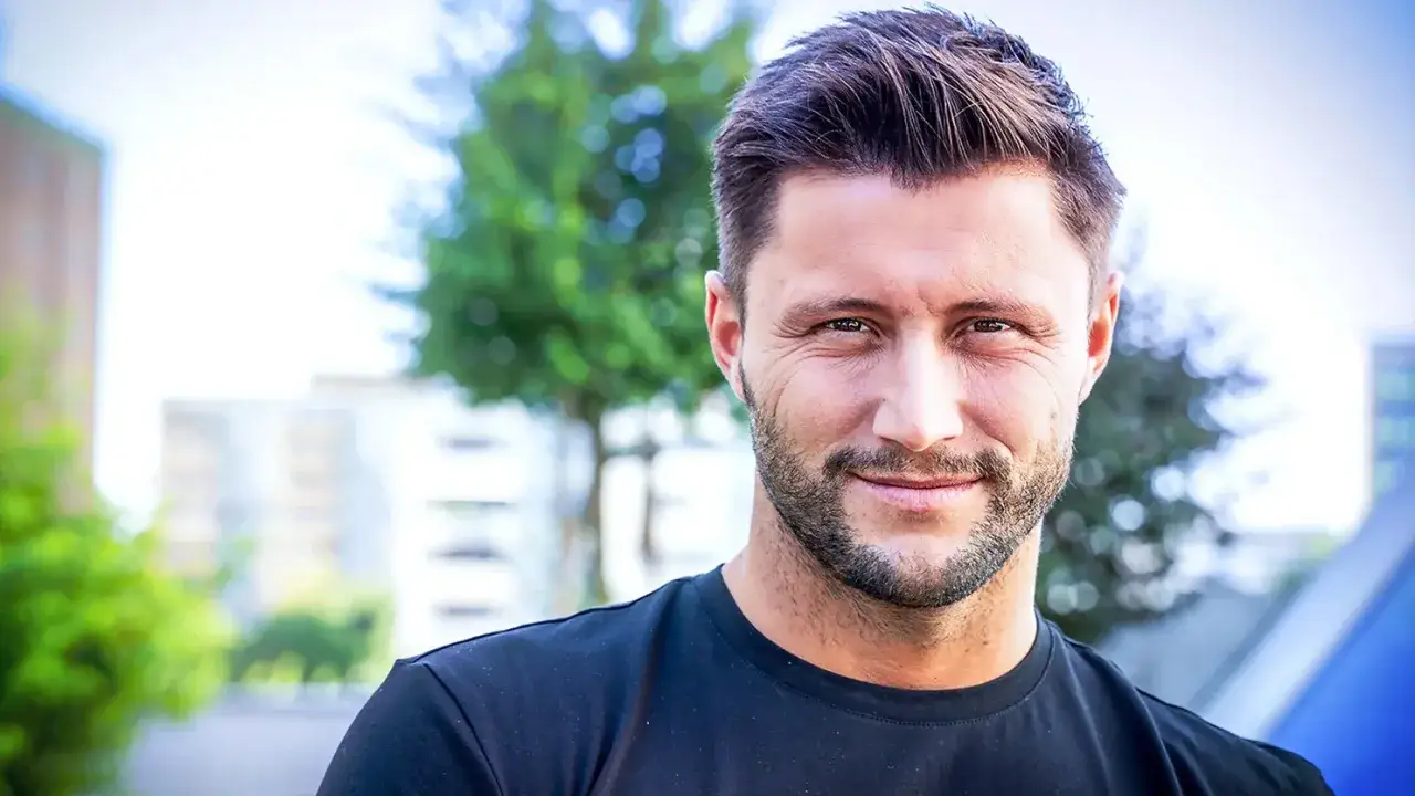 Uśmiechnięty Piotr Mróz w czarnej koszulce, zarośnięty, na tle zieleni i budynków.