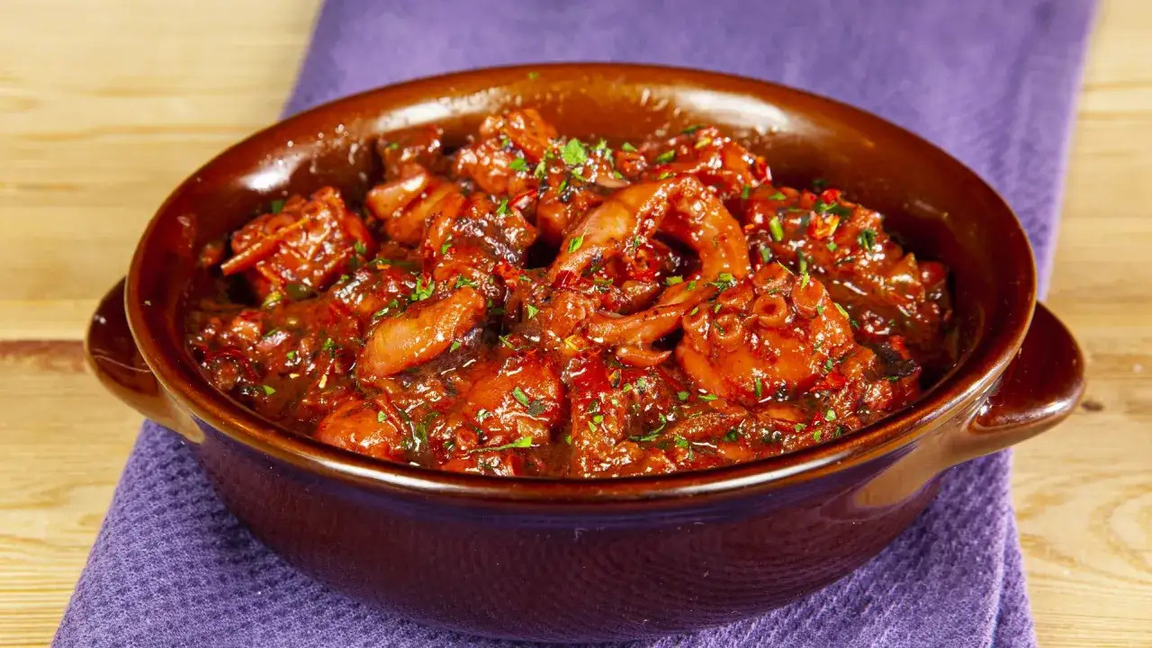 Un saporito polpo in umido, ricco di pomodoro e erbe aromatiche, servito in una ciotola di terracotta su un tovagliolo viola.