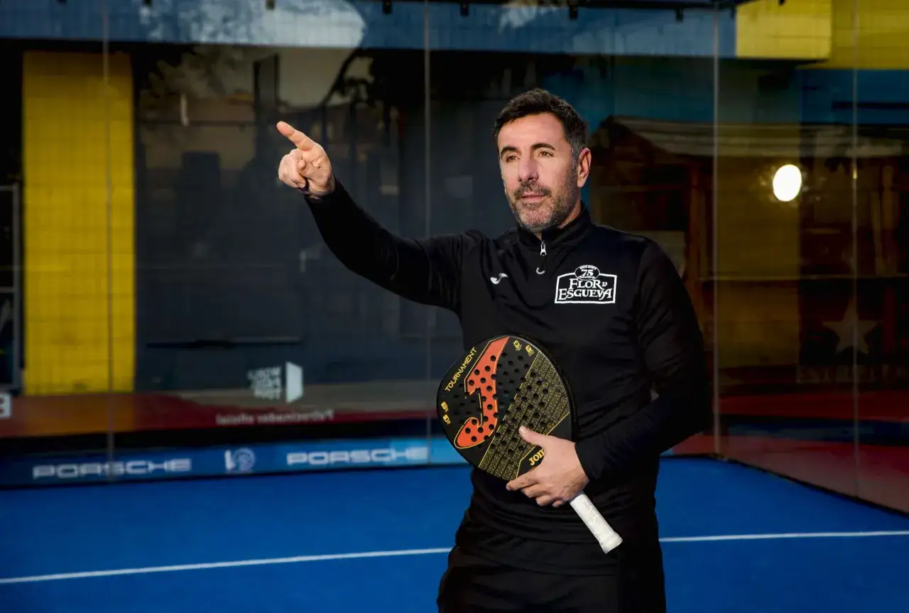 Gustavo Pratto, con una racchetta da padel in mano, indica con il dito verso l'alto su un campo da padel blu.