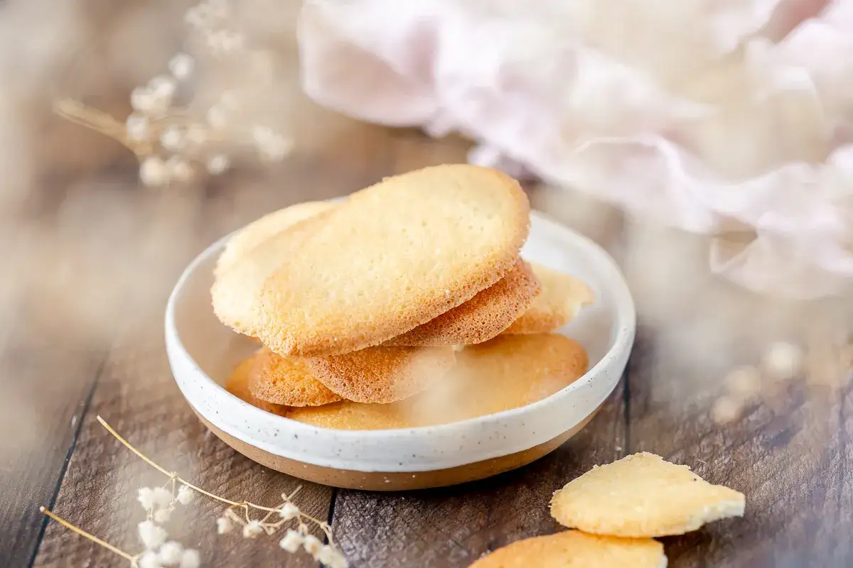 Delicate lingue di gatto dorate in una ciotola, su un tavolo di legno rustico, con un tessuto rosa sullo sfondo.