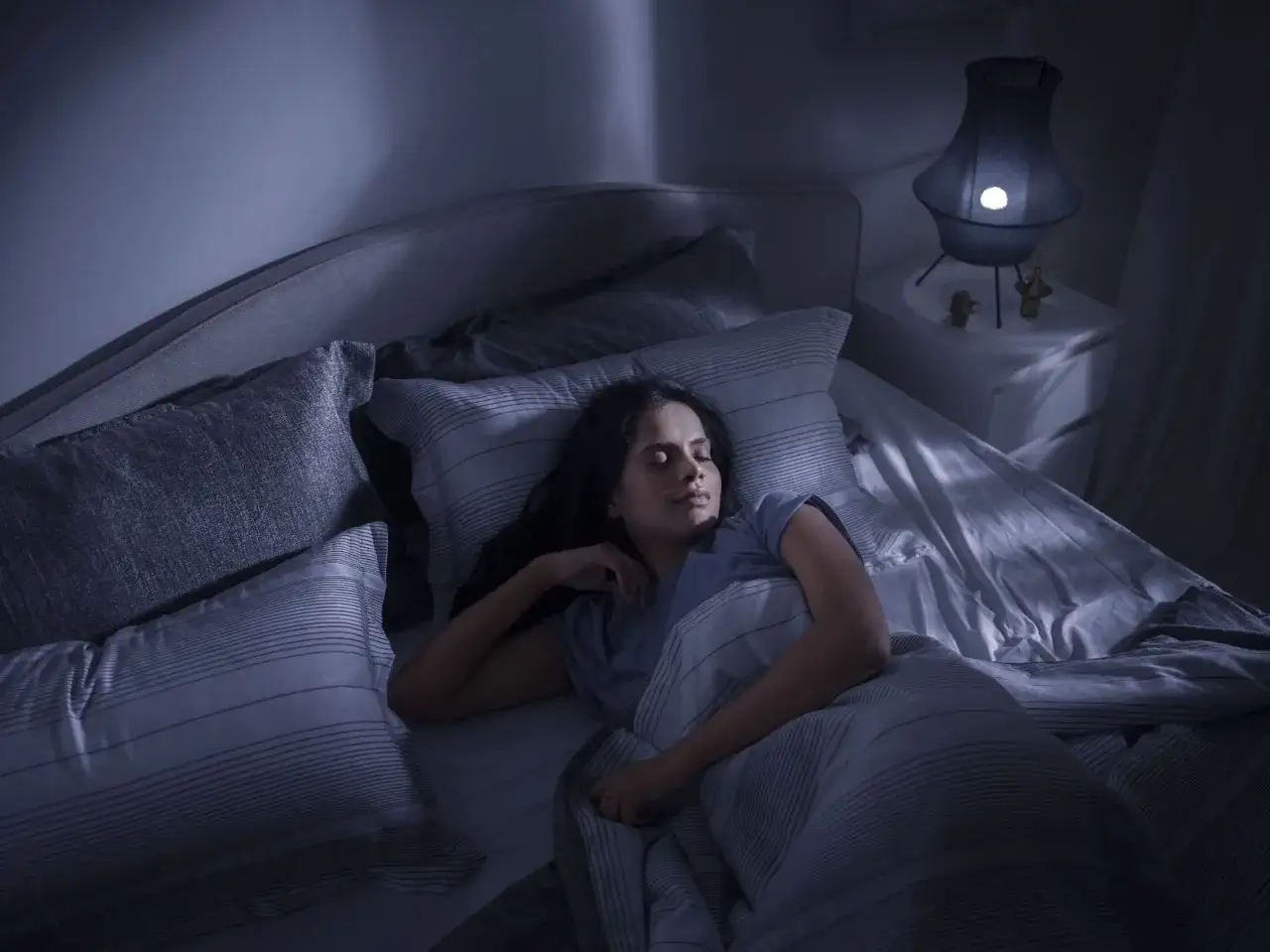 Eine Frau schl&auml;ft tief und fest in einem gem&uuml;tlichen Bett. Ihr Gesicht ist entspannt, und sie scheint einen **leichten Schlaf** zu genie&szlig;en, w&auml;hrend das sanfte Licht einer Nachttischlampe den Raum erhellt.