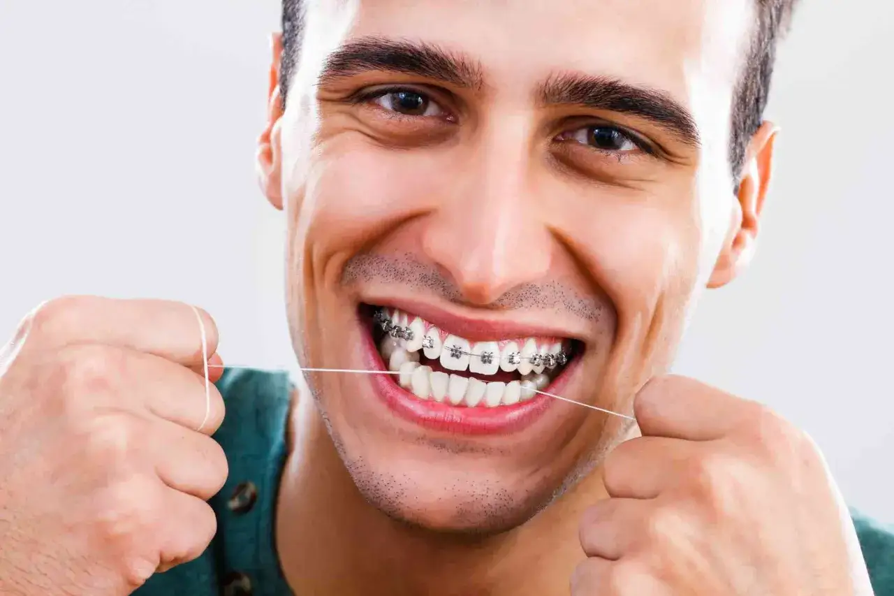 Uśmiechnięty mężczyzna z aparatem stałym czyści zęby nicią dentystyczną. Dbanie o higienę jest kluczowe przy noszeniu aparatu.