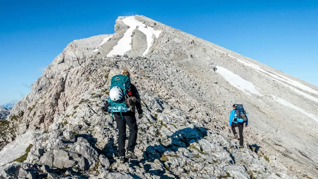 Zwei Wanderer steigen auf felsigem Untergrund den Watzmann empor. Schneefelder s&auml;umen den Weg zum Gipfel.