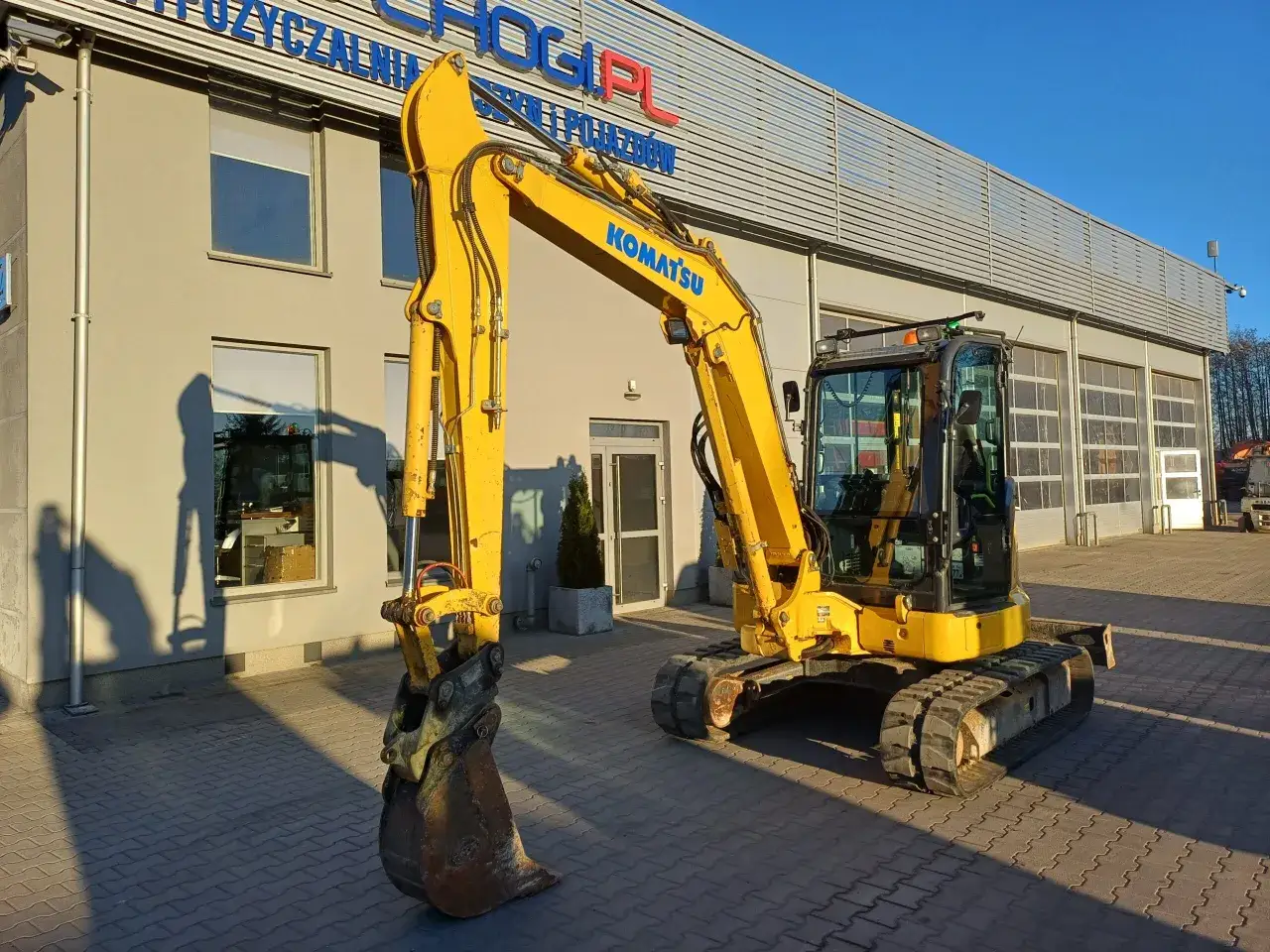 Żółta koparka Komatsu stoi przed budynkiem. Zastanawiasz się, ile bierze koparka za godzinę?
