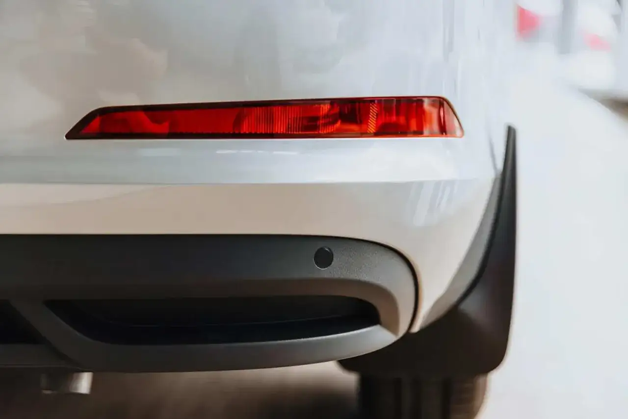 Vue rapprochée de l'arrière d'une voiture blanche, montrant un feu arrière rouge et un capteur de stationnement noir, des dispositifs réfléchissants pour la sécurité.