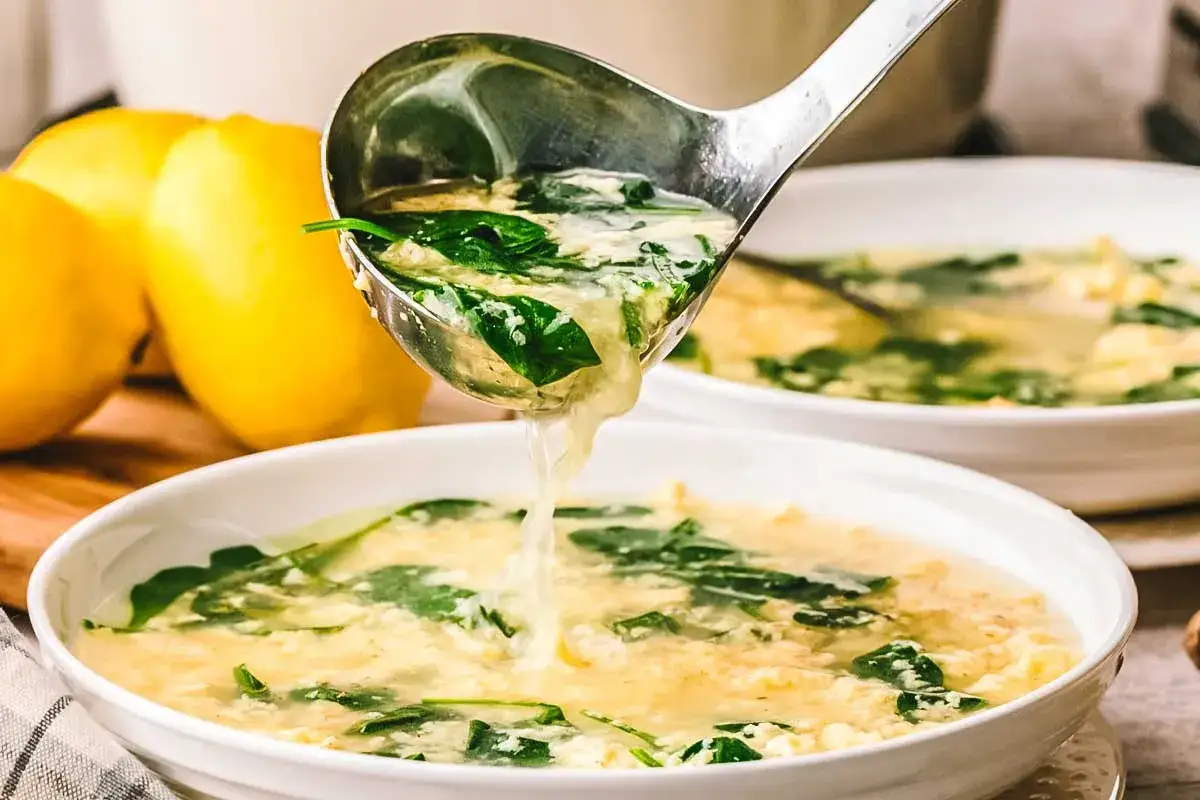 Chochelka nalewa do miski zupę stracciatella z listkami szpinaku. Obok leżą cytryny.