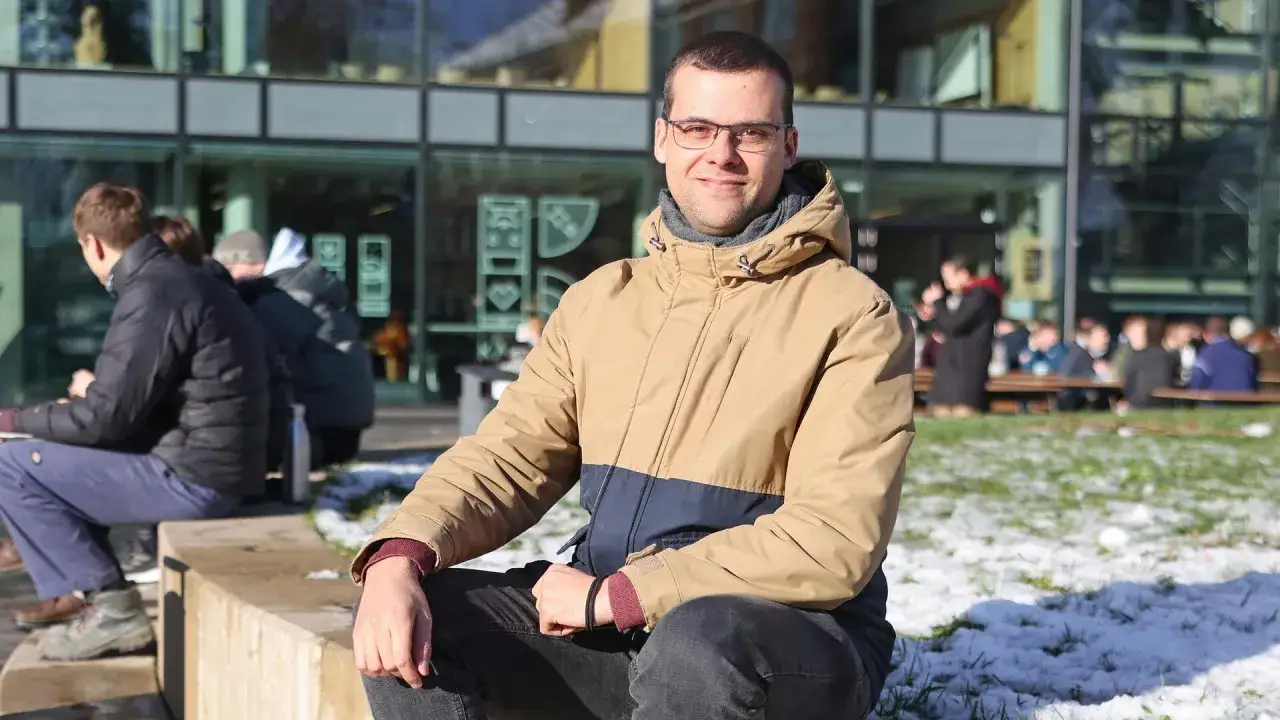 Ein Mann mit Brille sitzt im Freien vor einem modernen Gebäude. Er trägt eine zweifarbige Jacke und blickt lächelnd in die Kamera. Im Hintergrund sind andere Menschen zu sehen, die auf Bänken sitzen. Ein Bild, das einen **schwarm for future**-Moment ei...