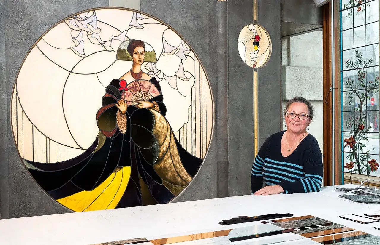 Un vitrail circulaire représente une femme élégante tenant un éventail. À côté, une artiste sourit, entourée de matériaux pour vitraux.