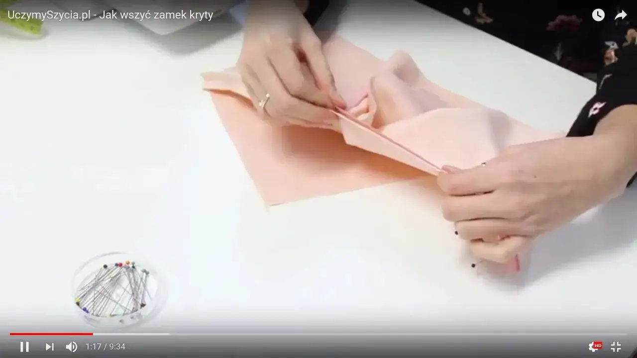 Ręce przygotowują materiał do szycia, by pokazać, jak wszyć zamek kryty. Szpilki czekają w pojemniczku.