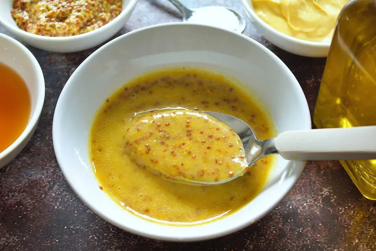 Cuchara con salsa miel y mostaza, lista para aderezar. Ingredientes a la vista: mostaza en grano, miel y aceite.