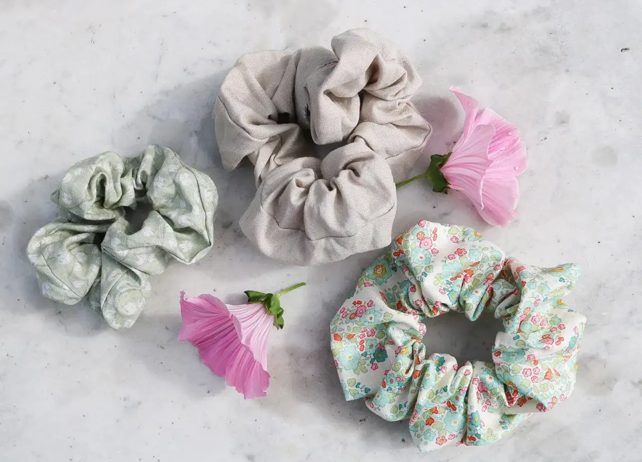 Tre sy scrunchies i olika mönster och färger, omgivna av rosa blommor på en marmoryta.