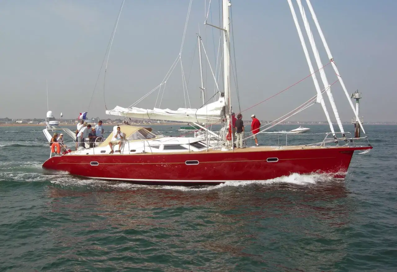 Un voilier rouge vif navigue sur l'eau, ses voiles blanches déployées. À son bord, plusieurs personnes profitent du soleil. Un ovni 25, peut-être un drone, survole la scène.