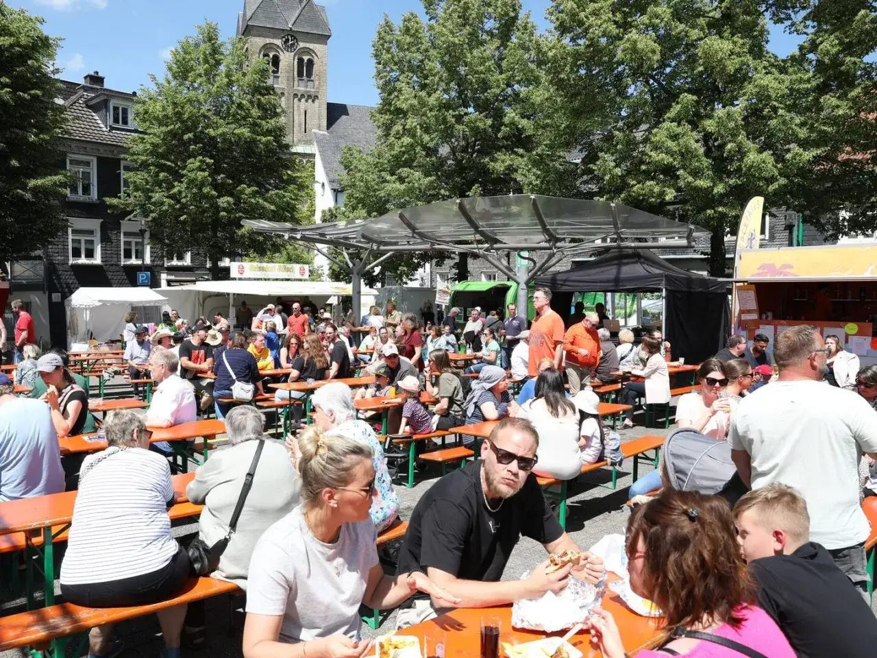Menschen genie&szlig;en Essen und Trinken bei sommerlichen Burscheid Veranstaltungen. Ein Kirchturm ragt im Hintergrund auf.