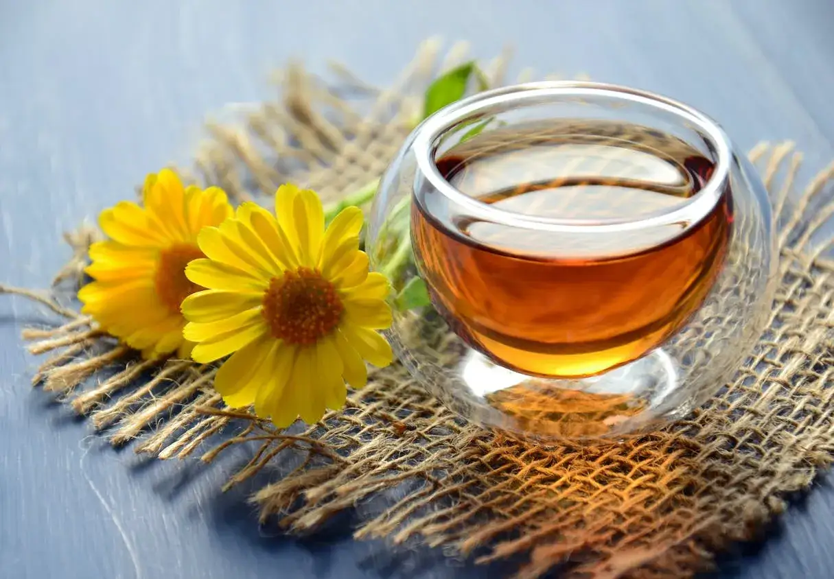 Zwei gelbe Bl&uuml;ten neben einem Glas Tee. Welcher Tee treibt nicht? Dieser goldene Aufguss, vielleicht aus Calendula, wirkt beruhigend.