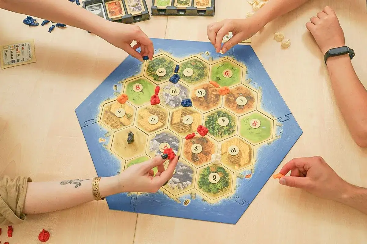 Gracze budują osady na planszy Catan. Widać fragmenty instrukcji i karty.