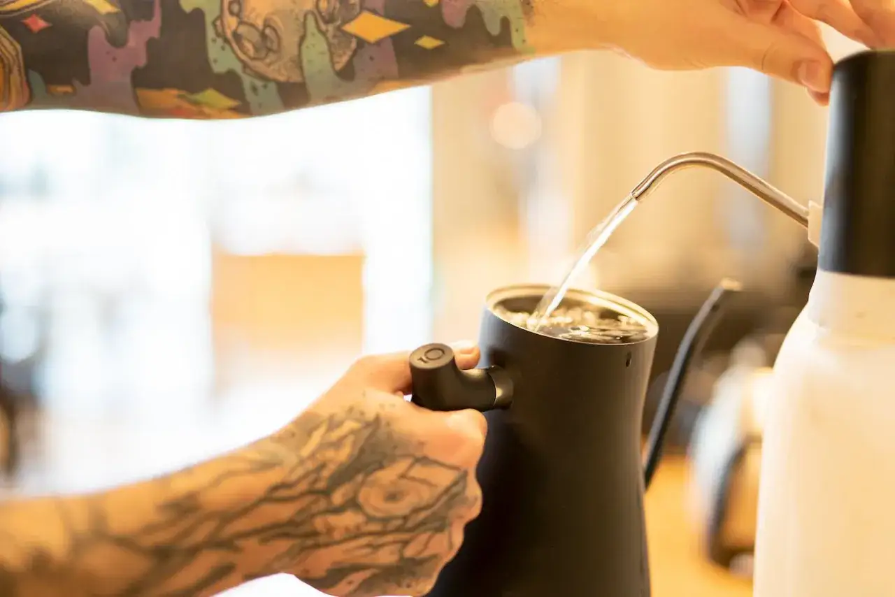 Tätowierte Hand gießt heißes Wasser in eine Kanne, bereit für die Zubereitung von Espresso.