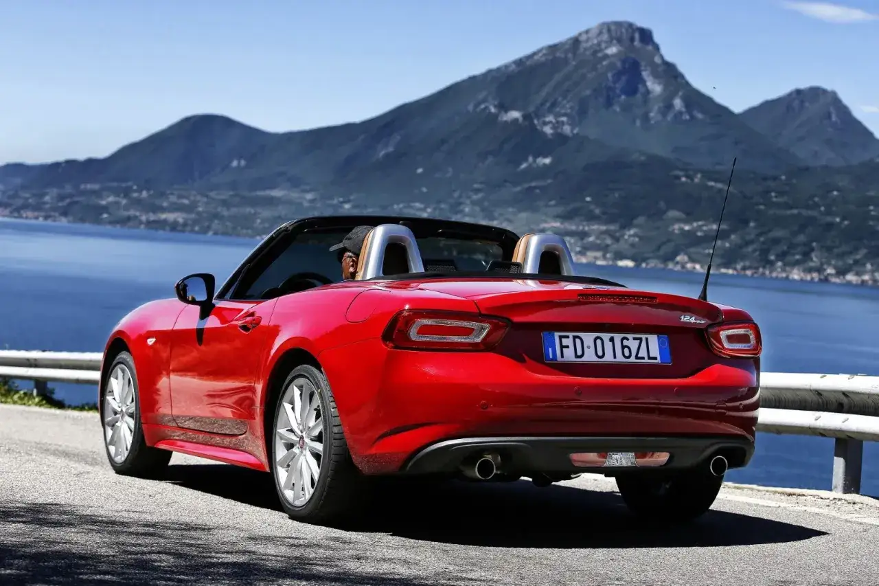 Czerwony Fiat 124 Spider z opuszczonym dachem jedzie malowniczą drogą nad jeziorem, z górami w tle.
