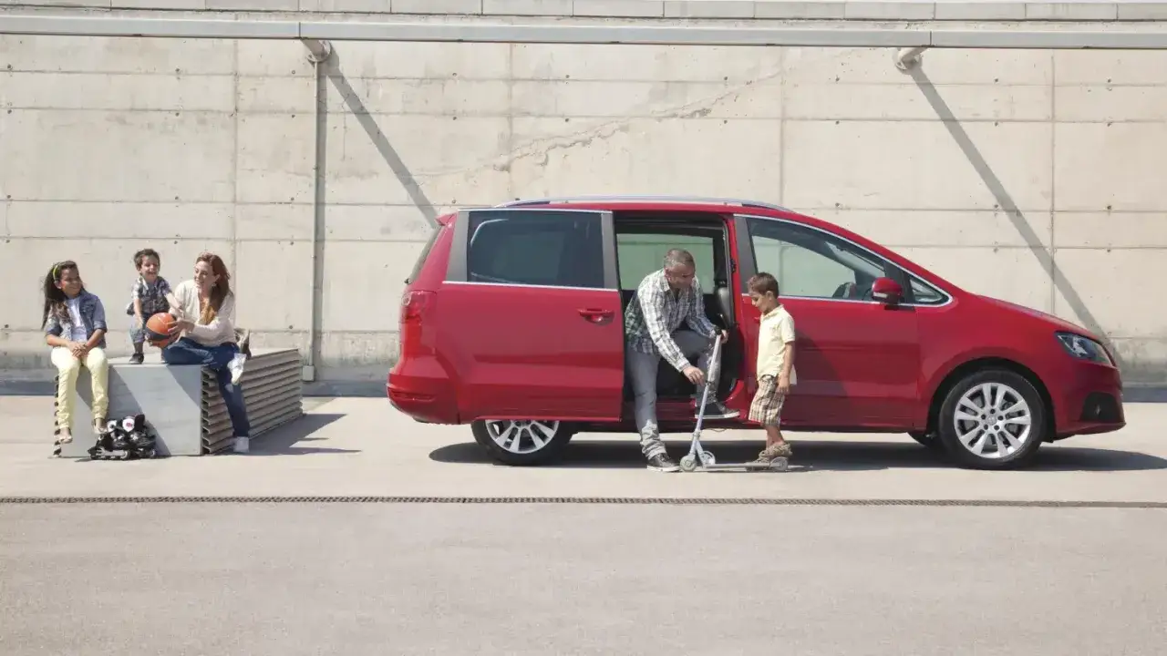 Ein rotes Auto mit 3 Kindern. Ein Vater steigt mit seinem Sohn aus dem Auto, w&auml;hrend die Mutter mit zwei Kindern auf einer Bank sitzt.