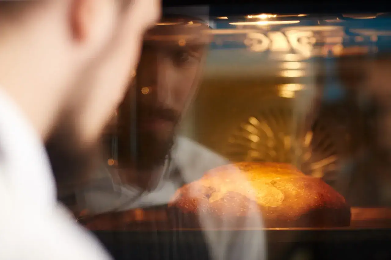 Ein Mann mit Bart schaut in den Ofen, wo ein Brot bäckt. Für knuspriges Brot muss man beim Brot backen Wasser in den Ofen.