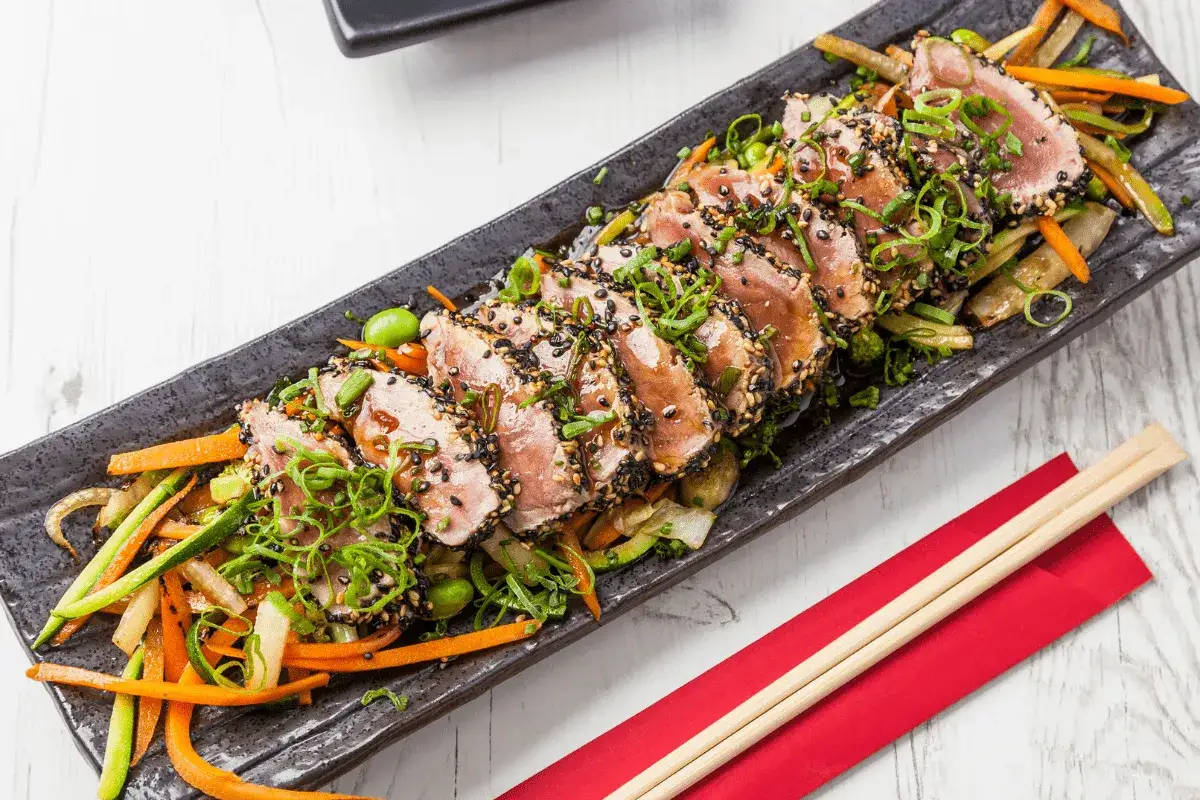Tataki de bonito con sésamo negro y verduras salteadas, servido en plato negro con palillos.