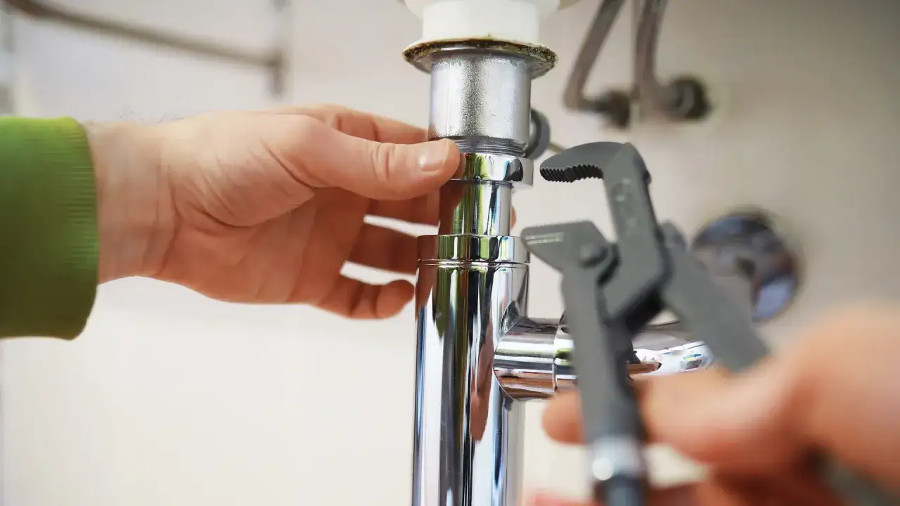 Montaż zaworów wody pod zlewem. Ręce majsterkowicza dokręcają chromowane elementy instalacji hydraulicznej kluczem.