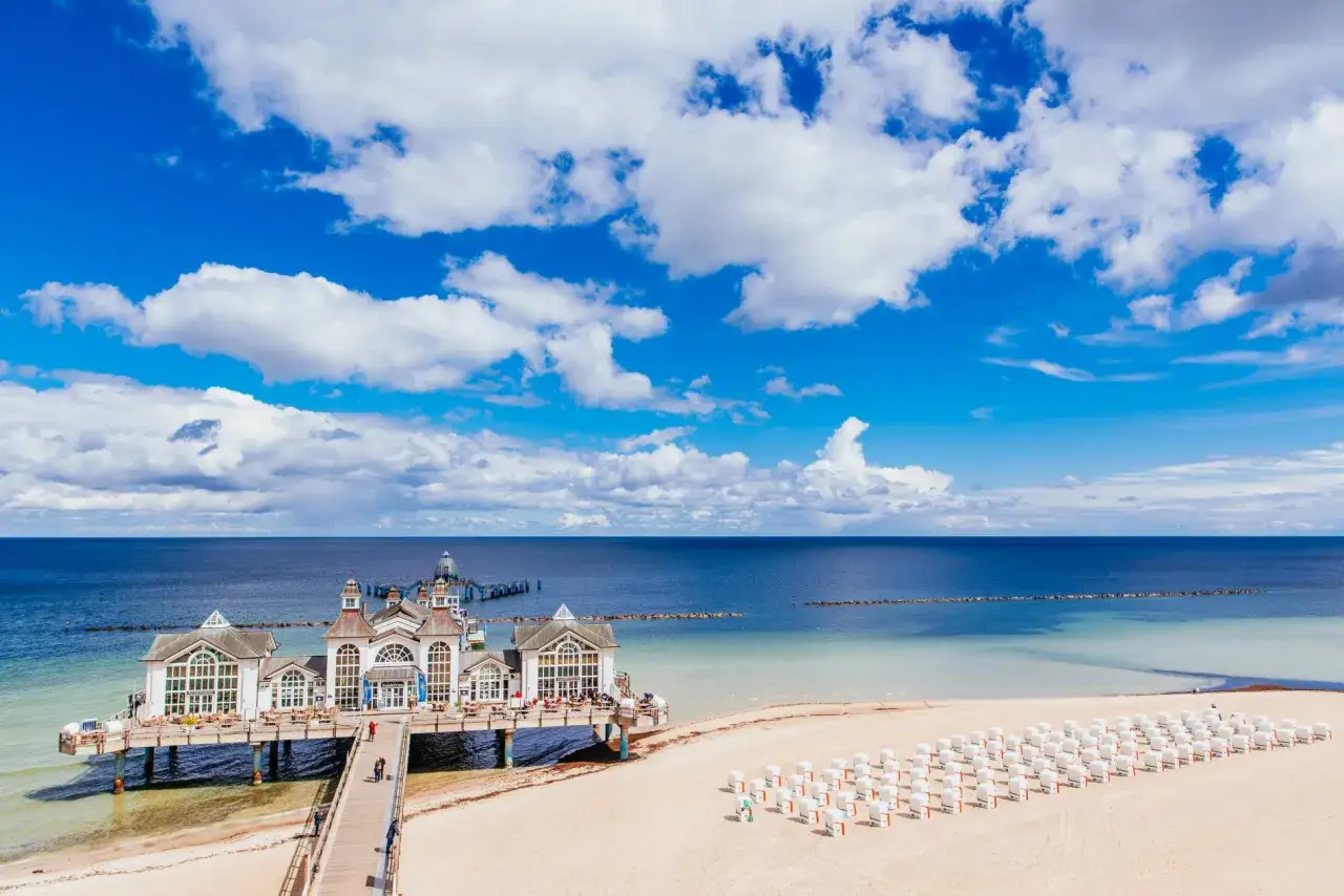 Ein sonniger Tag im Ostseebad Sellin. Die Seebrücke mit ihren weißen Gebäuden ragt ins Meer, daneben reihen sich Strandkörbe am Sandstrand.
