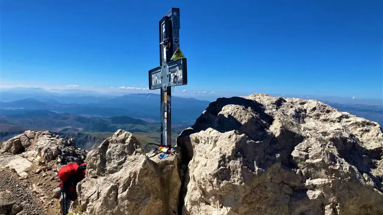 Cima rocciosa con croce metallica, zaino rosso e panorama mozzafiato. Il **corno grande** domina il paesaggio.