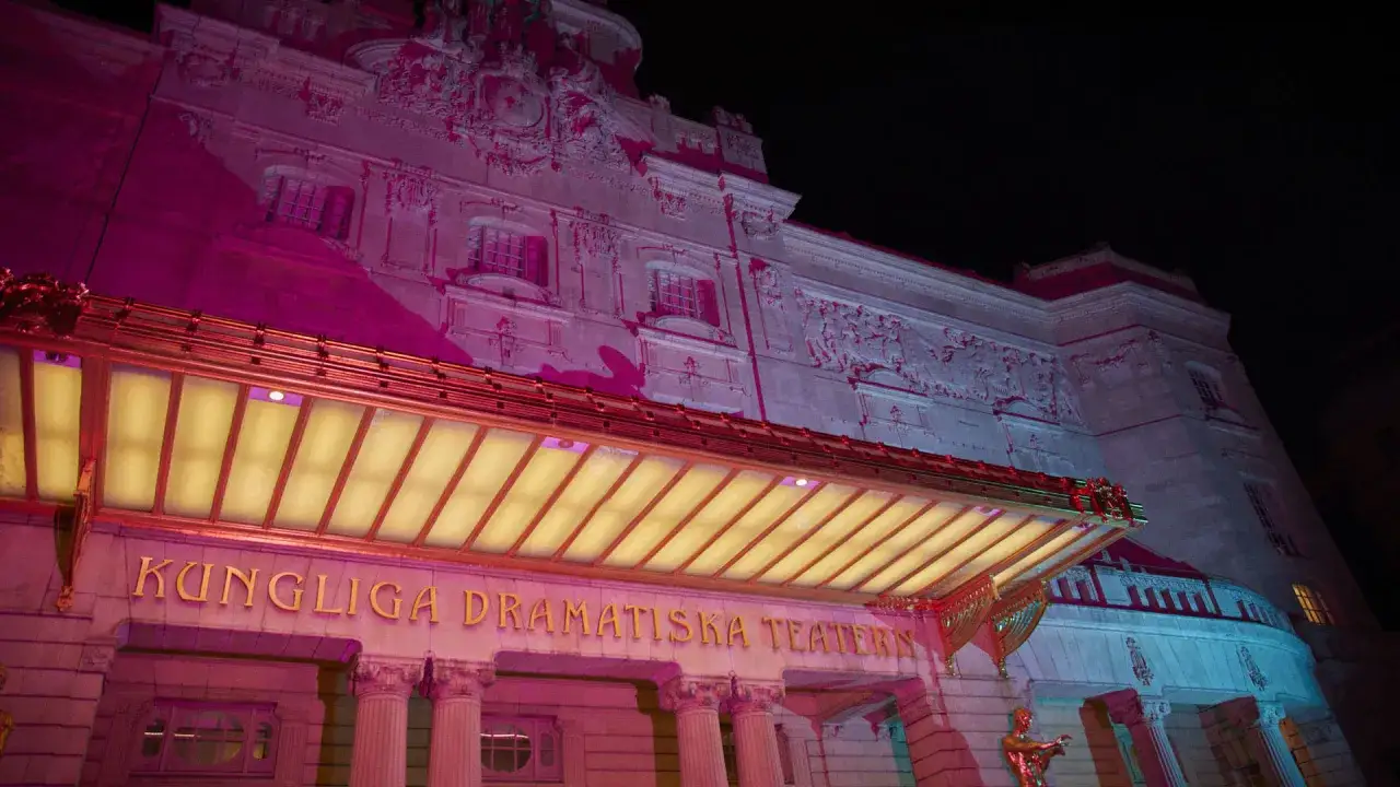 Kungliga Dramatiska Teatern badar i rosa och blått ljus. Den storslagna fasaden med sina detaljerade skulpturer och gyllene entré lyser upp natten.