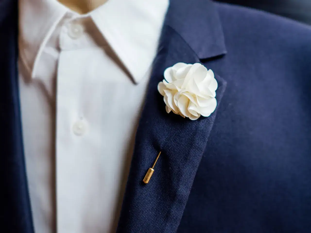 Elegancka, biała butonierka w kształcie kwiatu ozdabia granatową marynarkę. Złoty agrafka dodaje szyku.