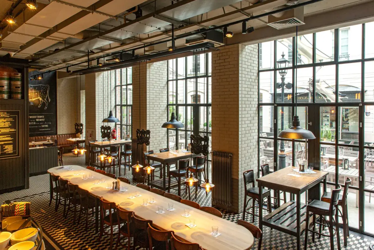 Wnętrze restauracji z długim stołem i krzesłami, okna z widokiem na ulicę. Na ścianie rysunek wołu, jak w 