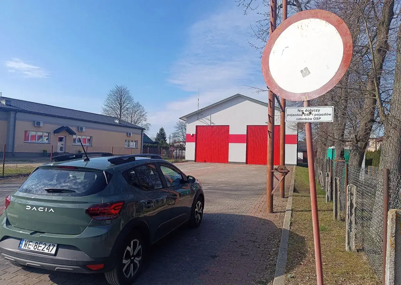 Zielony Dacia Sandero zaparkowany przy remizie OSP. Znak zakazu zbliżania się do garaży, by uniknąć mandatu za parkowanie.