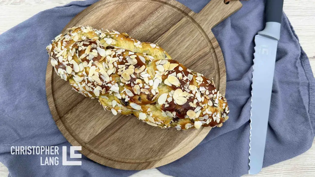 Ein goldbrauner Hefezopf mit Rosinen, bestreut mit Mandelblättchen, liegt auf einem Holzbrett. Daneben ein Messer.
