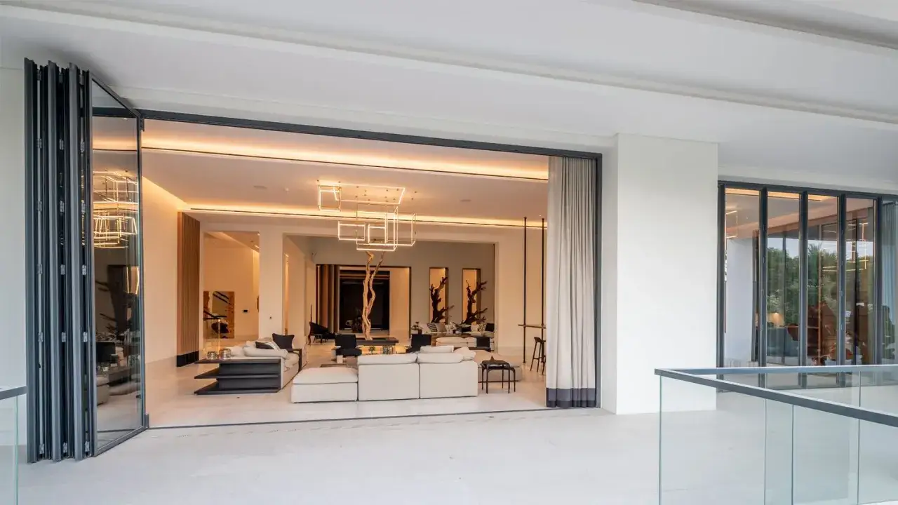 Amplio sal&oacute;n moderno con sof&aacute;s blancos, una l&aacute;mpara geom&eacute;trica y una puerta abatible que conecta con el exterior.