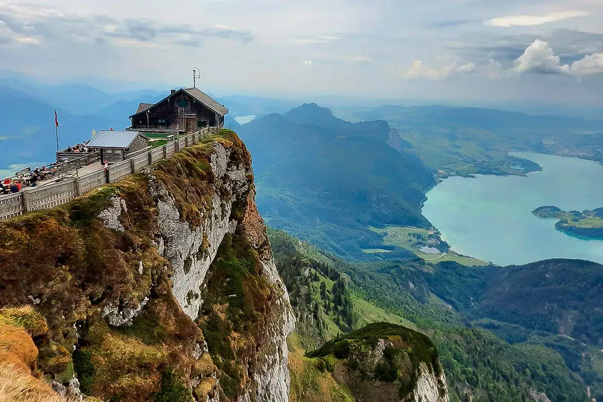 Ein Gasthaus thront auf einem Felsvorsprung am Himmelspforte Schafberg. Der Blick schweift &uuml;ber einen t&uuml;rkisfarbenen See und gr&uuml;ne Berge.