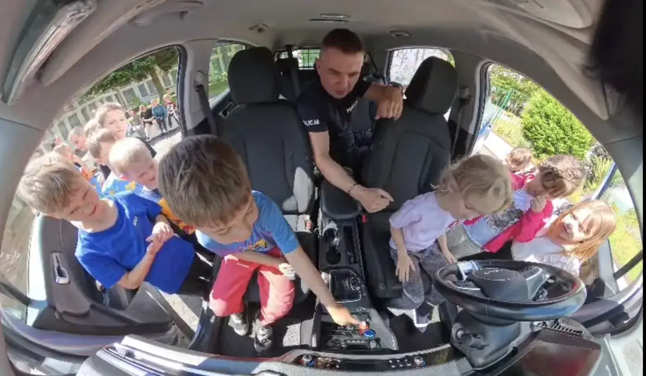 Policjant w radiowozie z dziećmi. Wesoła atmosfera, żadnego utrudniania kontakt&oacute;w z dzieckiem.