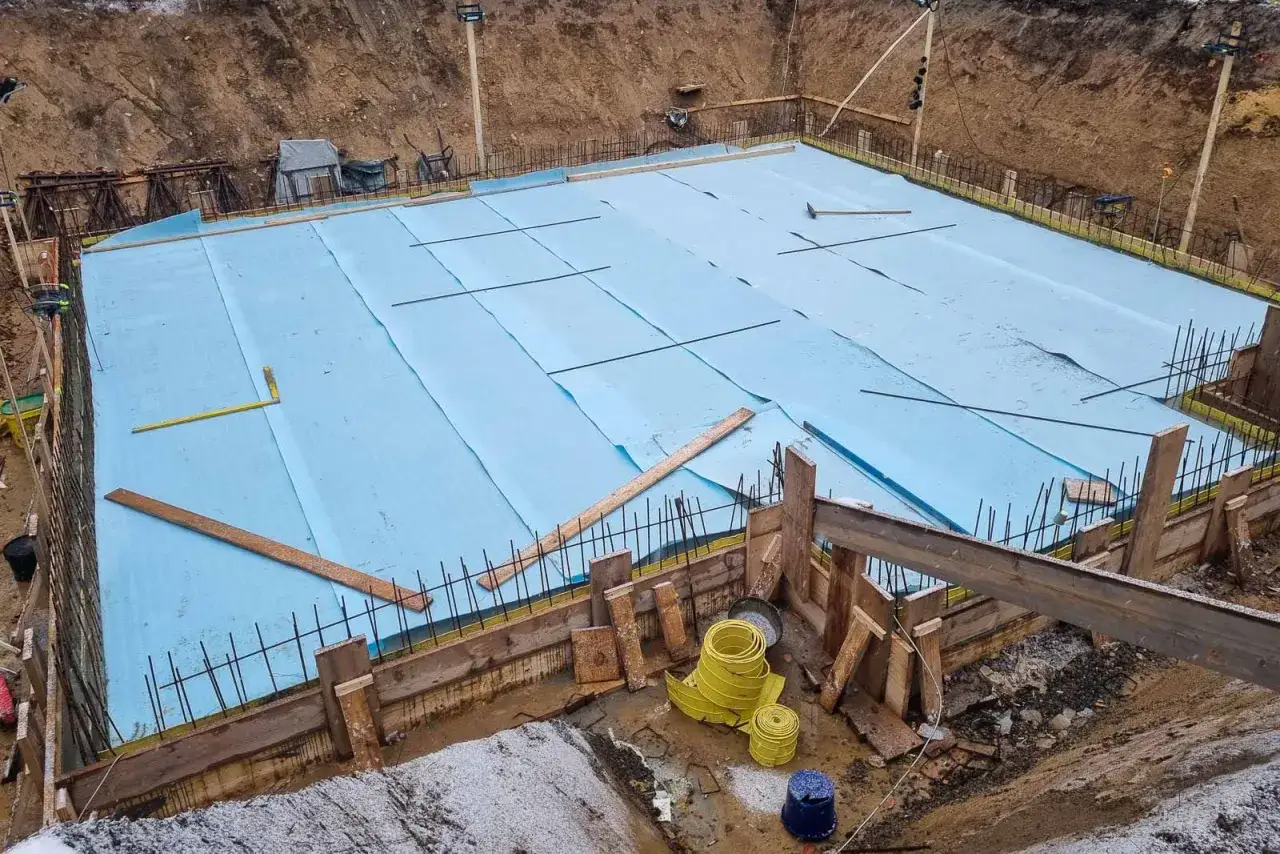 Bauarbeiten im Regen: Eine Baustelle mit blauer Folie und Bewehrung, bereit für das Betonieren.