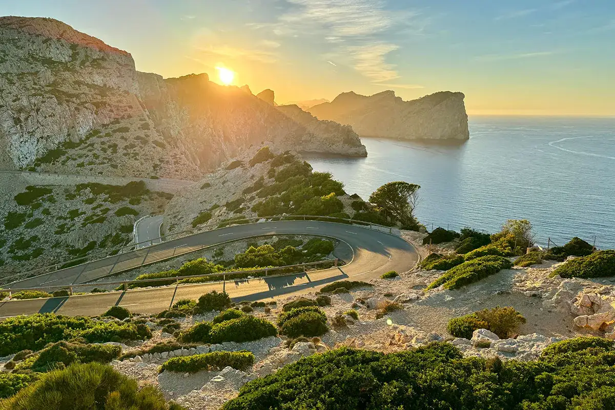 Serpentinenstraße auf Mallorca im Dezember, die sich durch felsige Küstenlandschaft schlängelt. Die Sonne wirft lange Schatten auf die Straße und das Meer.