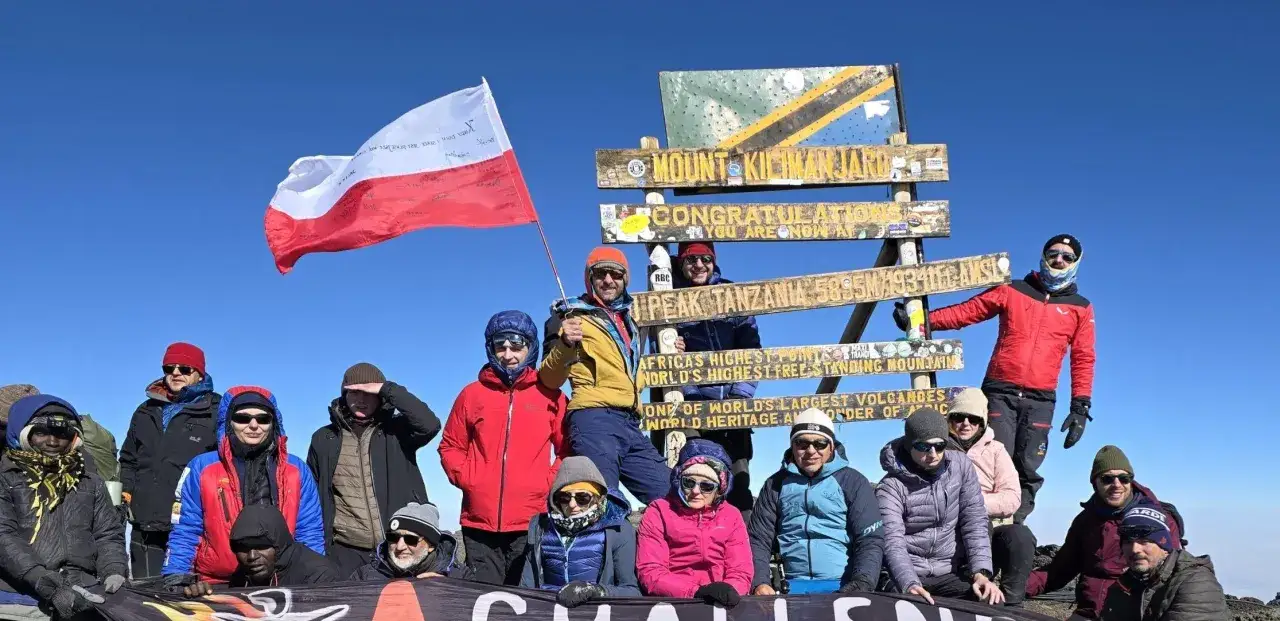 Grupa zdobywców na szczycie Kilimandżaro z polską flagą. Wszyscy uśmiechnięci, gotowi na dalszą podróż, pokazując, jak dobrać tempo karawany do najsłabszego uczestnika wyprawy.