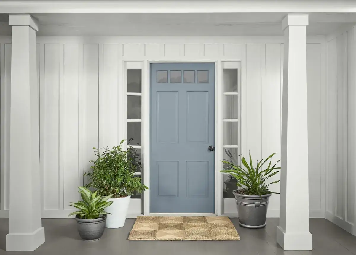 Puerta de madera exterior pintada de azul, flanqueada por plantas y pilares blancos. Un felpudo rústico añade calidez.