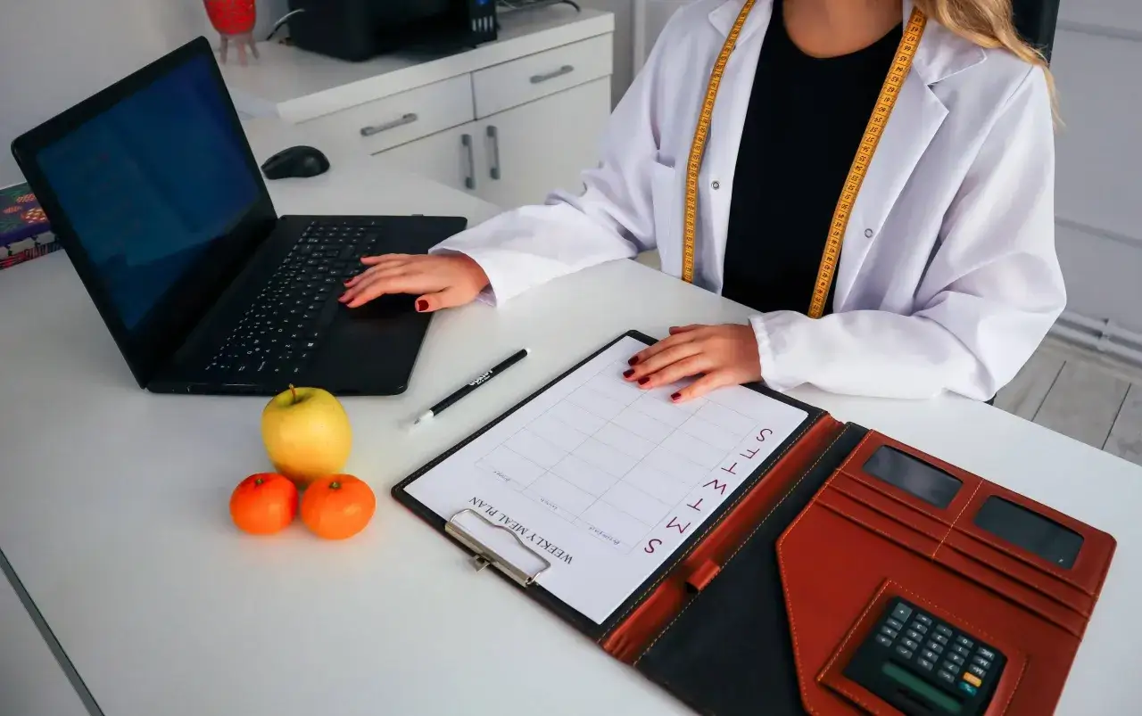 Nutricionista en su escritorio con plan semanal, frutas y calculadora. Averigua cuanto cobra un nutricionista para tu plan de salud.