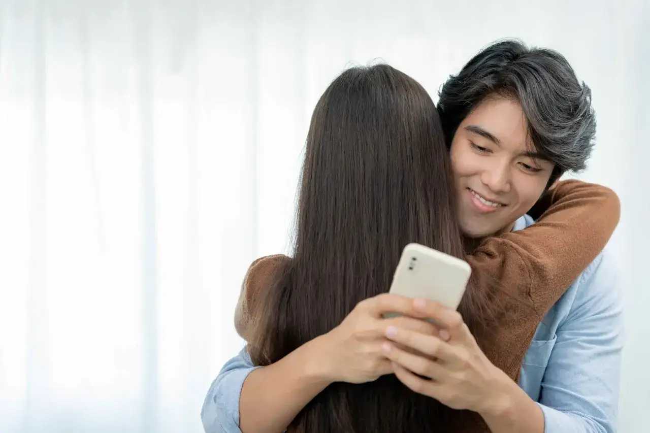 Un homme enlace une femme, son regard se porte sur son téléphone. Comment savoir si mon mari me trompe ? L'image suggère une possible inquiétude.