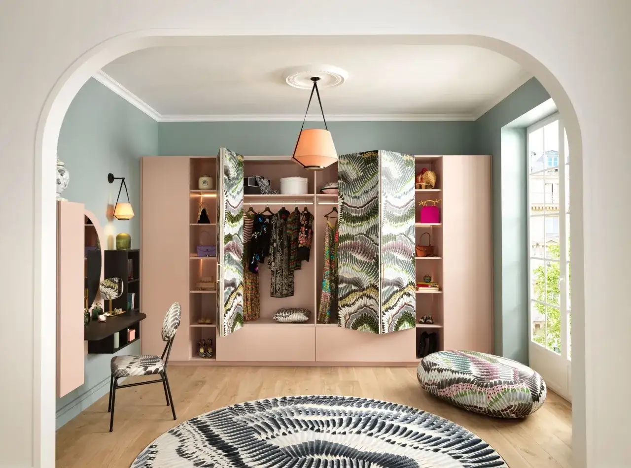 Un dressing rose pâle offre une grande profondeur, avec des étagères remplies de sacs et de chaussures. Des rideaux aux motifs audacieux cachent des vêtements.