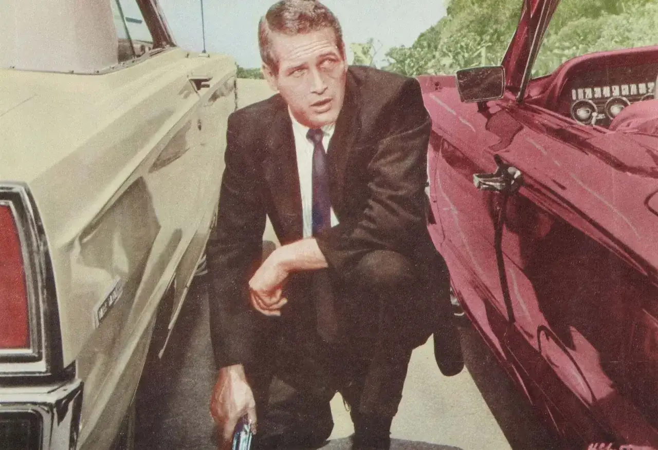 Paul Newman i kostym, hukandes mellan två bilar, en ljusgul och en mörkröd. En scen som påminner om hans ikoniska roller i filmer med Paul Newman.