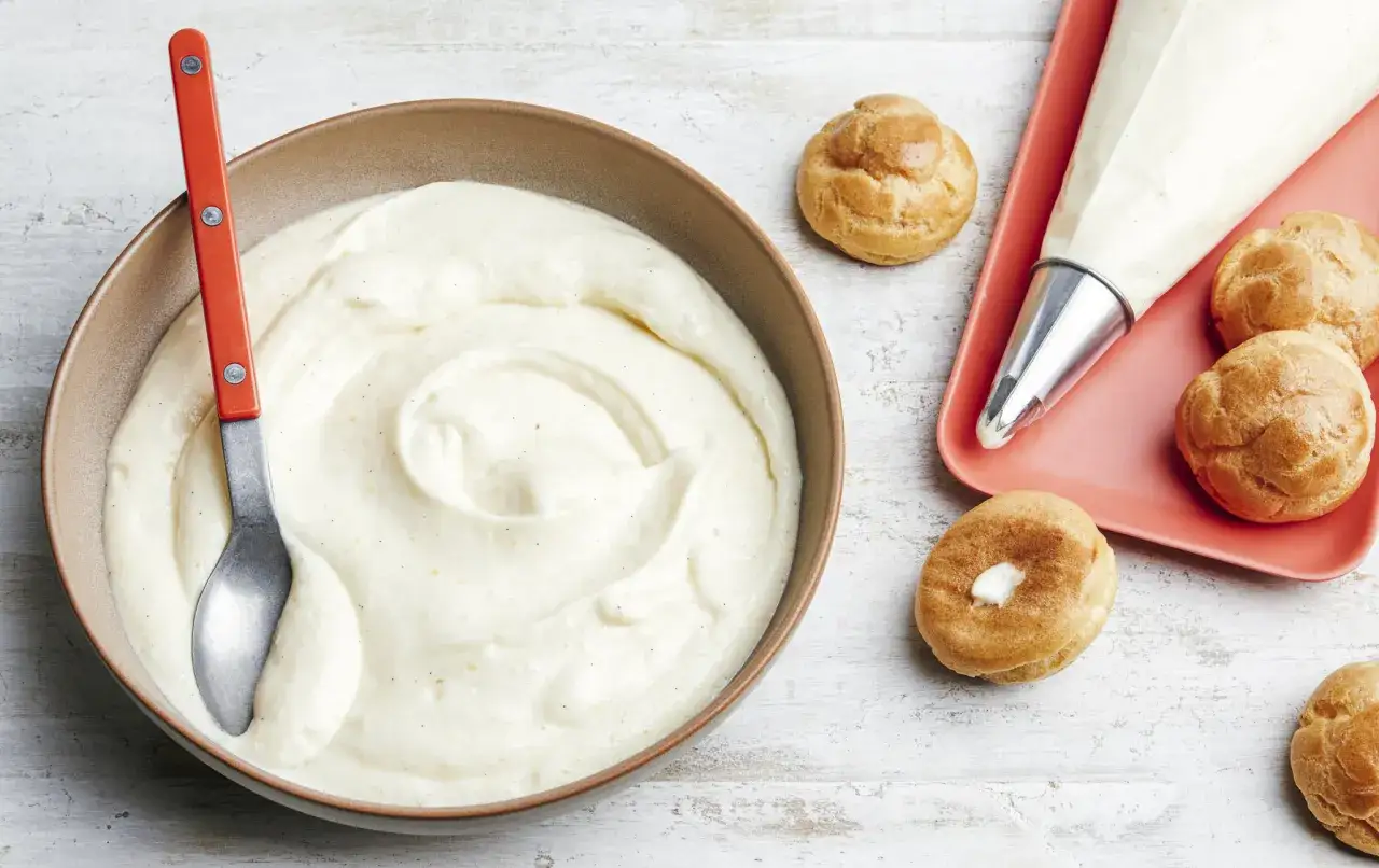 Un bol de crème chiboust onctueuse, une poche à douille et des petits choux prêts à être garnir.