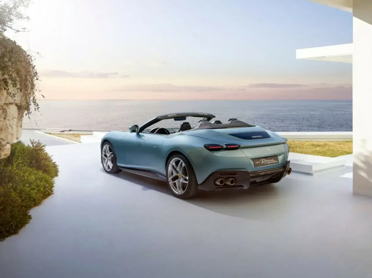 Błękitny roadster Ferrari Roma z otwartym dachem stoi na tarasie z widokiem na morze o zachodzie słońca.