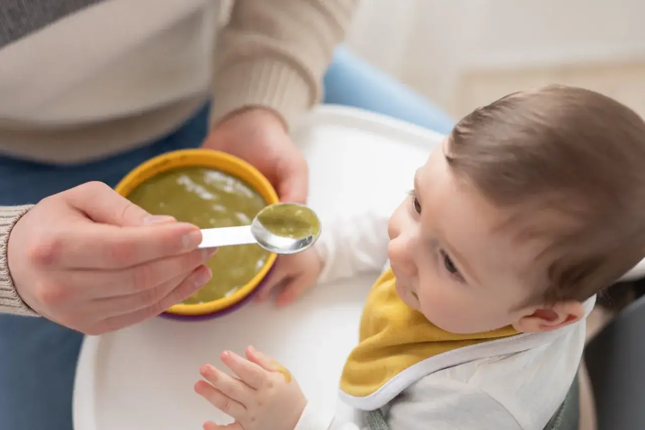Un bambino con un bavaglino giallo guarda con curiosit&agrave; un cucchiaino di pappa verde, un riflesso di estrusione che anticipa il primo assaggio.